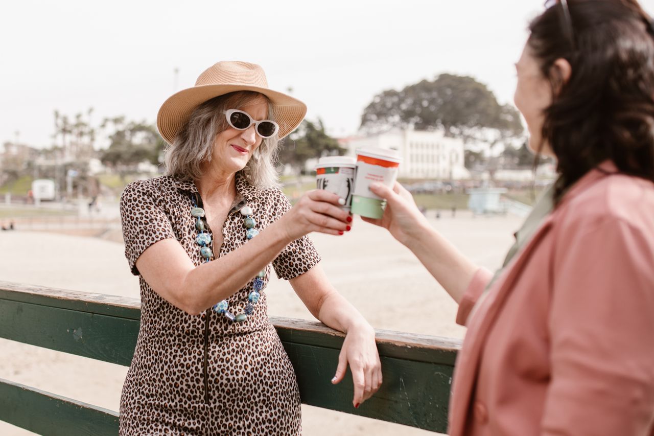 Two women enjoying a day outdoors, toasting with coffee in a sunny setting.