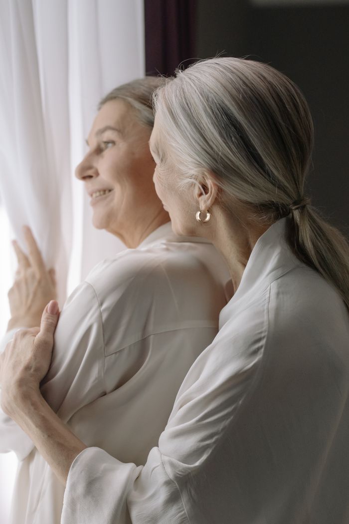 who-we-are Two senior women warmly embrace, enjoying a peaceful moment by a window.