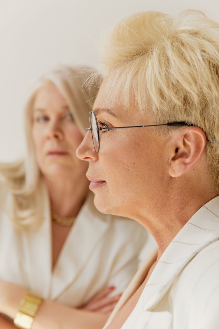 Close-up profile of two elegant mature women wearing business attire, showcasing eyeglasses fashion.