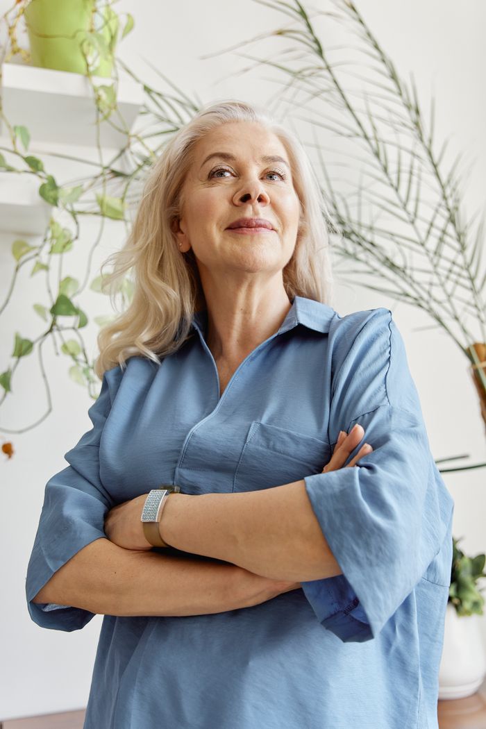 services-03 Portrait of a confident senior woman indoors, arms crossed, surrounded by indoor plants.