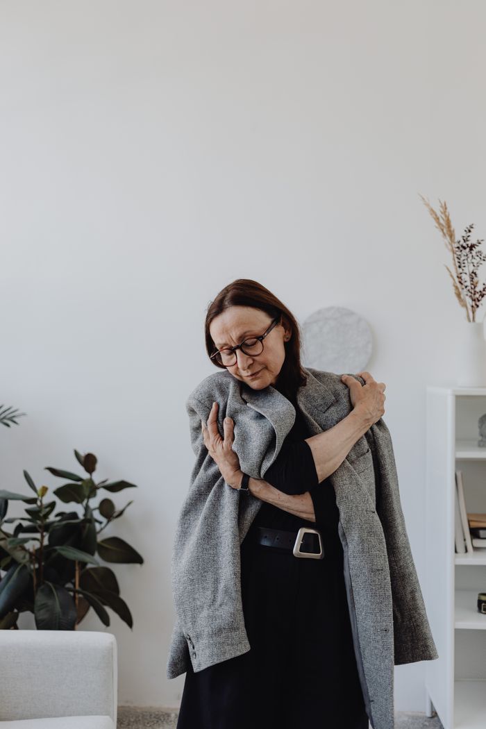 services-04 Senior woman indoors embracing gray coat, expressing introspective emotion.